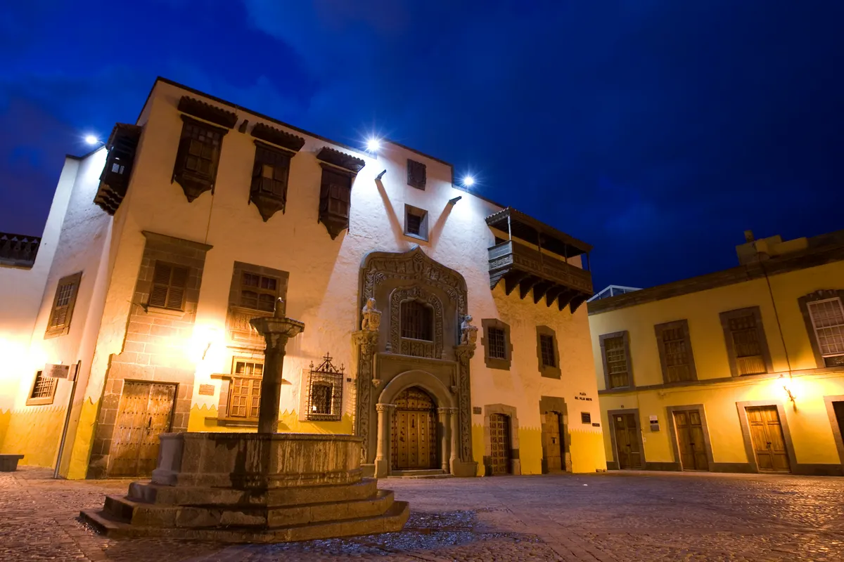 The illuminated façade of the Casa de Colón at dusk, with its carved stone doorway and wooden balconies