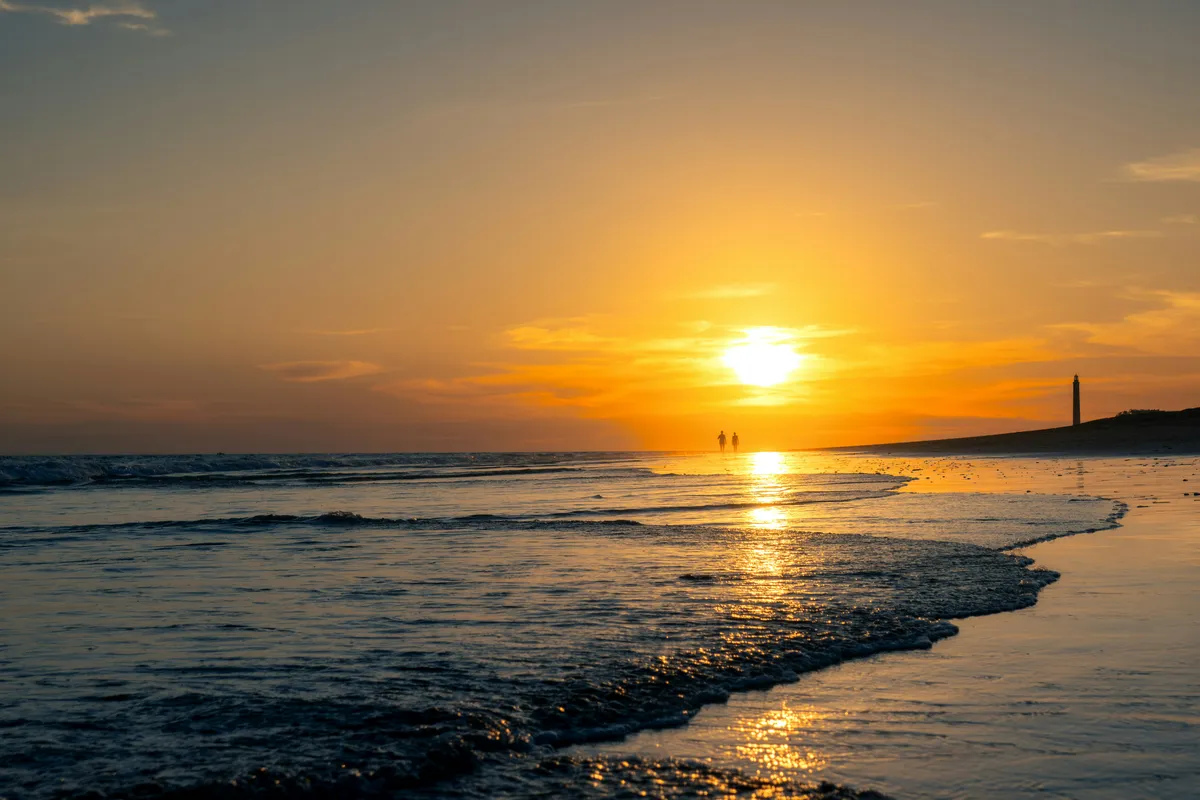 Puesta de sol en la orilla de la playa de Maspalomas, con el faro al fondo