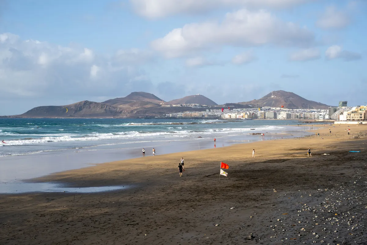 Playa de Las Canteras con olas y bandera roja, con la ciudad y los volcanes de La Isleta al fondo